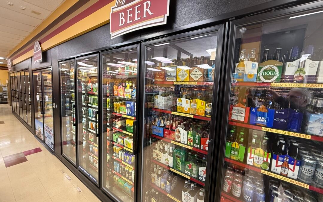 International beer cooler section at Salem Village Market featuring a wide selection of domestic and imported beer brands including Corona, Budweiser, Bud Light, Heineken, O'Doul's, and Dos Equis stocked across multiple glass-door refrigerators.